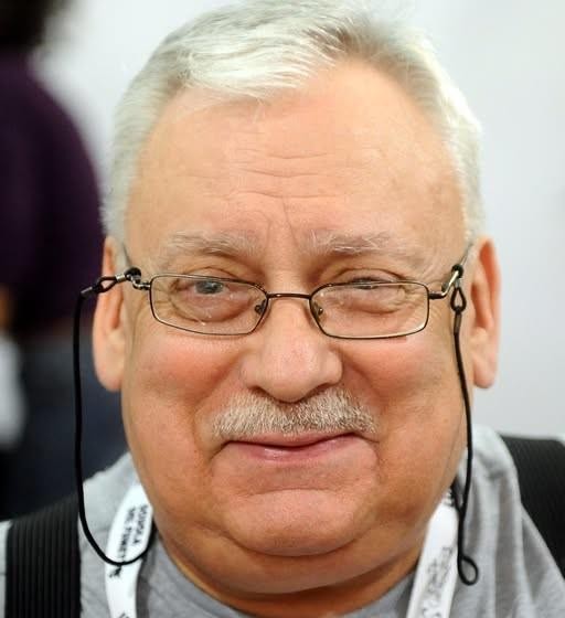 Zbiór książek Andrzeja Sapkowskiego, w tym Wiedźmin i Trylogia Husycka, ułożony na półce, z subtelnym, magicznym blaskiem.