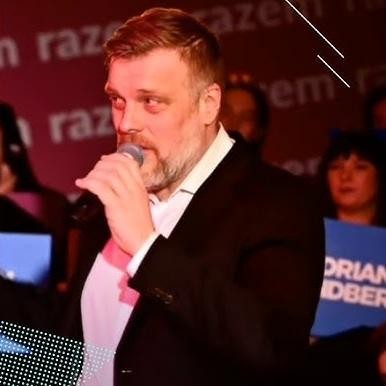 Adrian Zandberg podczas pełnego pasji wystąpienia publicznego, z gestykulacją podkreślającą jego zaangażowanie i przekonanie.