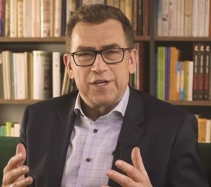 Maciej Orłoś prowadzący szkolenie z wystąpień publicznych, gestykulujący, w profesjonalnym otoczeniu.