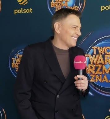 Krzysztof Ibisz w akcji na planie telewizyjnym, prezentujący program z mikrofonem w dłoni, z dynamicznym tłem studia TV.